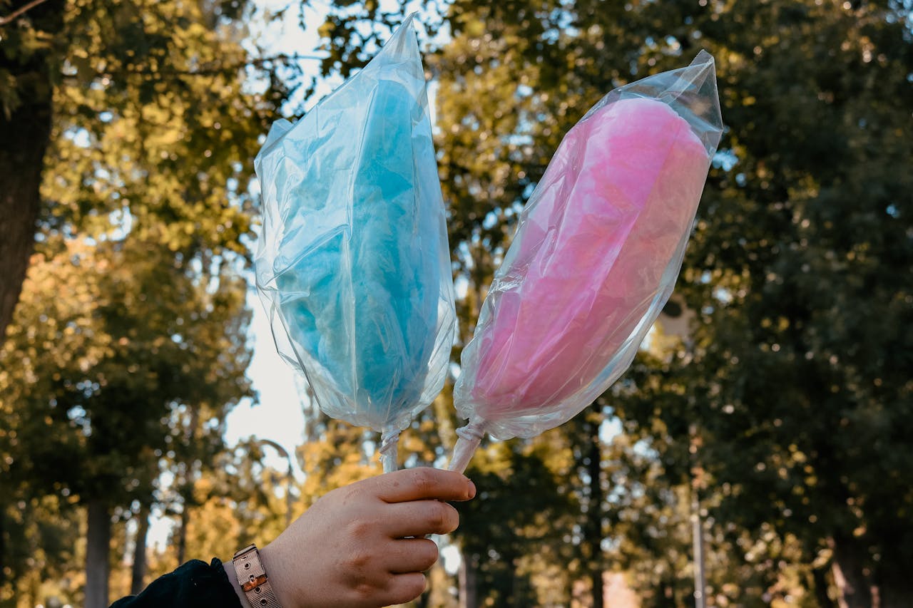 Two cotton candy sticks, one blue and one pink, held outdoors in a park setting.